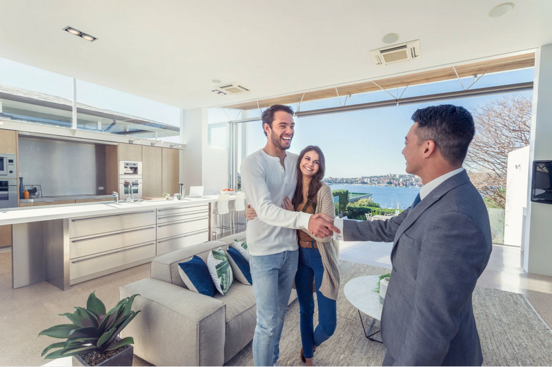 A real estate agent shakes hands with a couple in a modern, bright apartment with a scenic water view in the background.