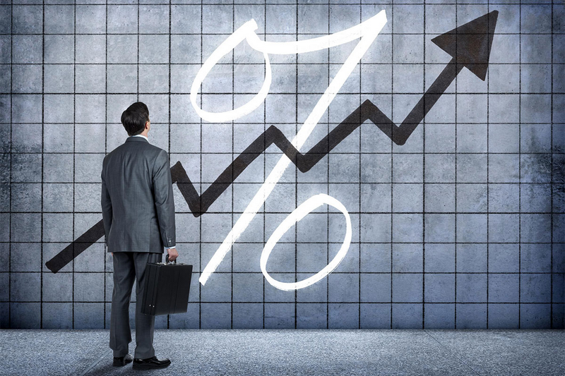 A businessman stands in front of a large percentage symbol with an upward arrow on a tiled wall.